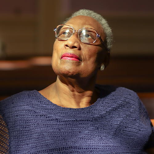 the image shows a black woman with short gray hair wearing glasses and pink lipstick looking off to her right, she is wearing a blue knit sweater