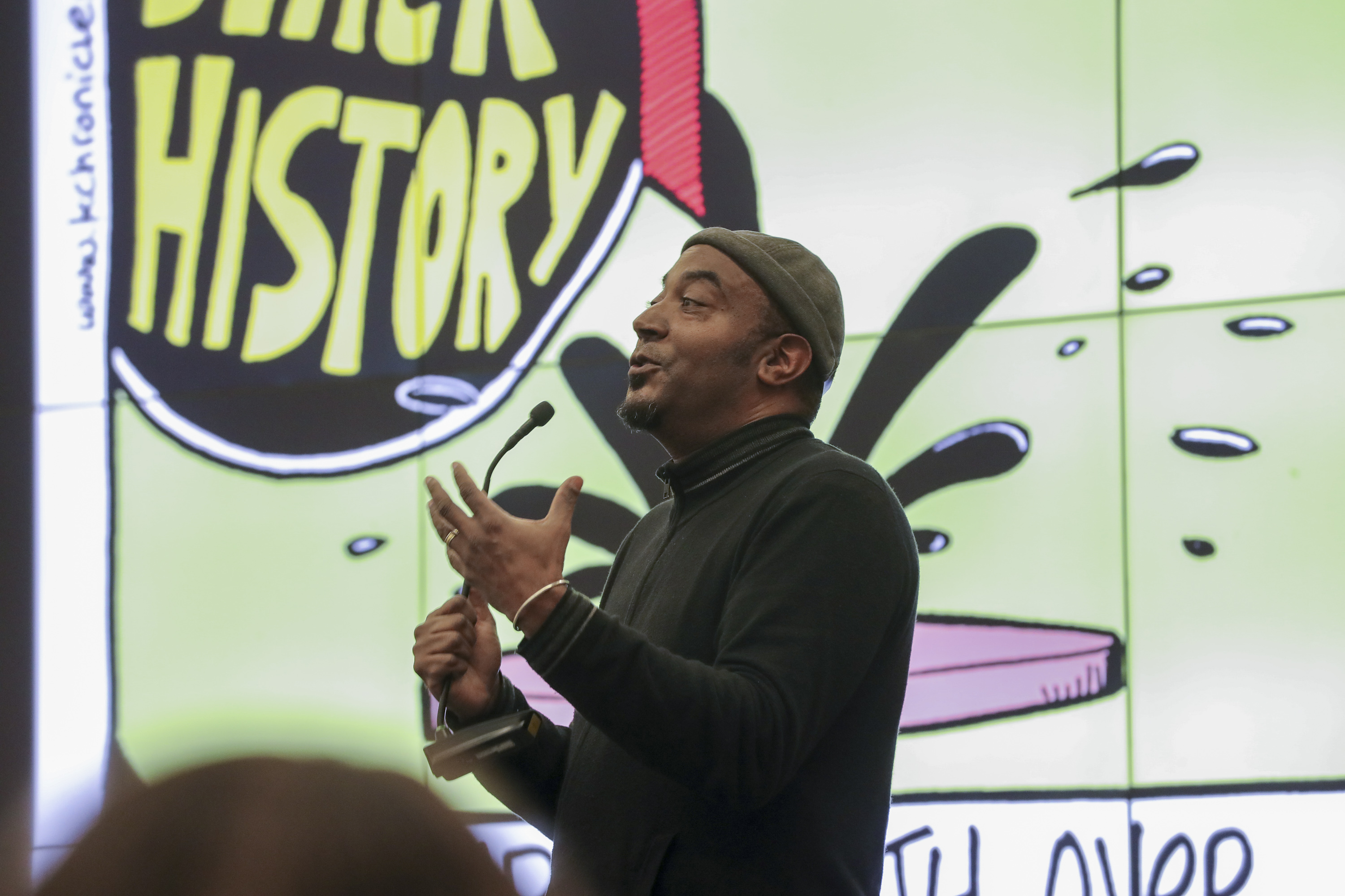 Cartoonist Keith Knight speaking in front of a screen showing some of his art at the annual Black History Month Lecture