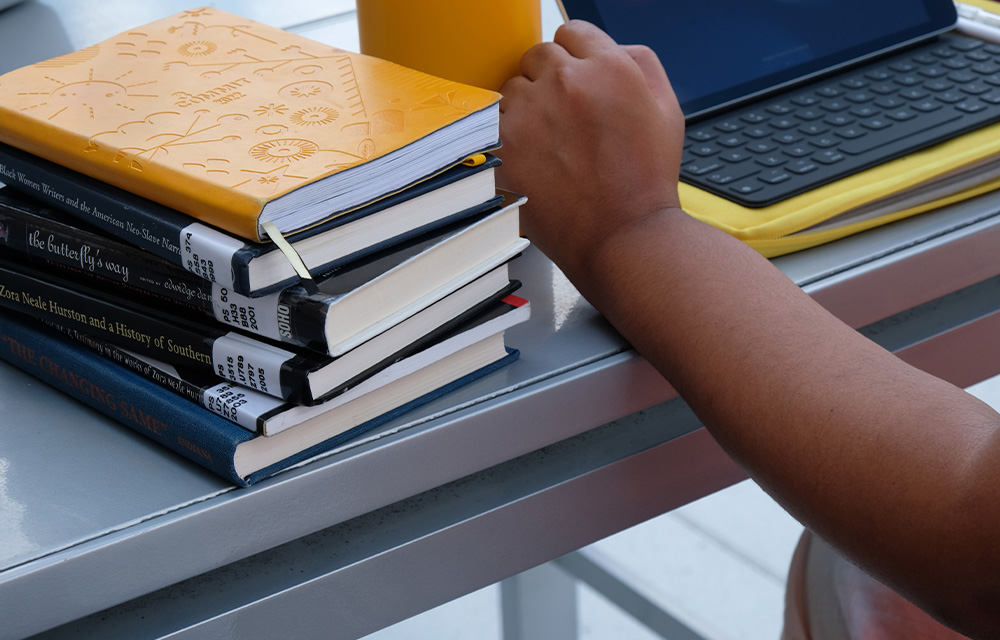 A stack of books next to a laptop, water bottle and a person's hand.