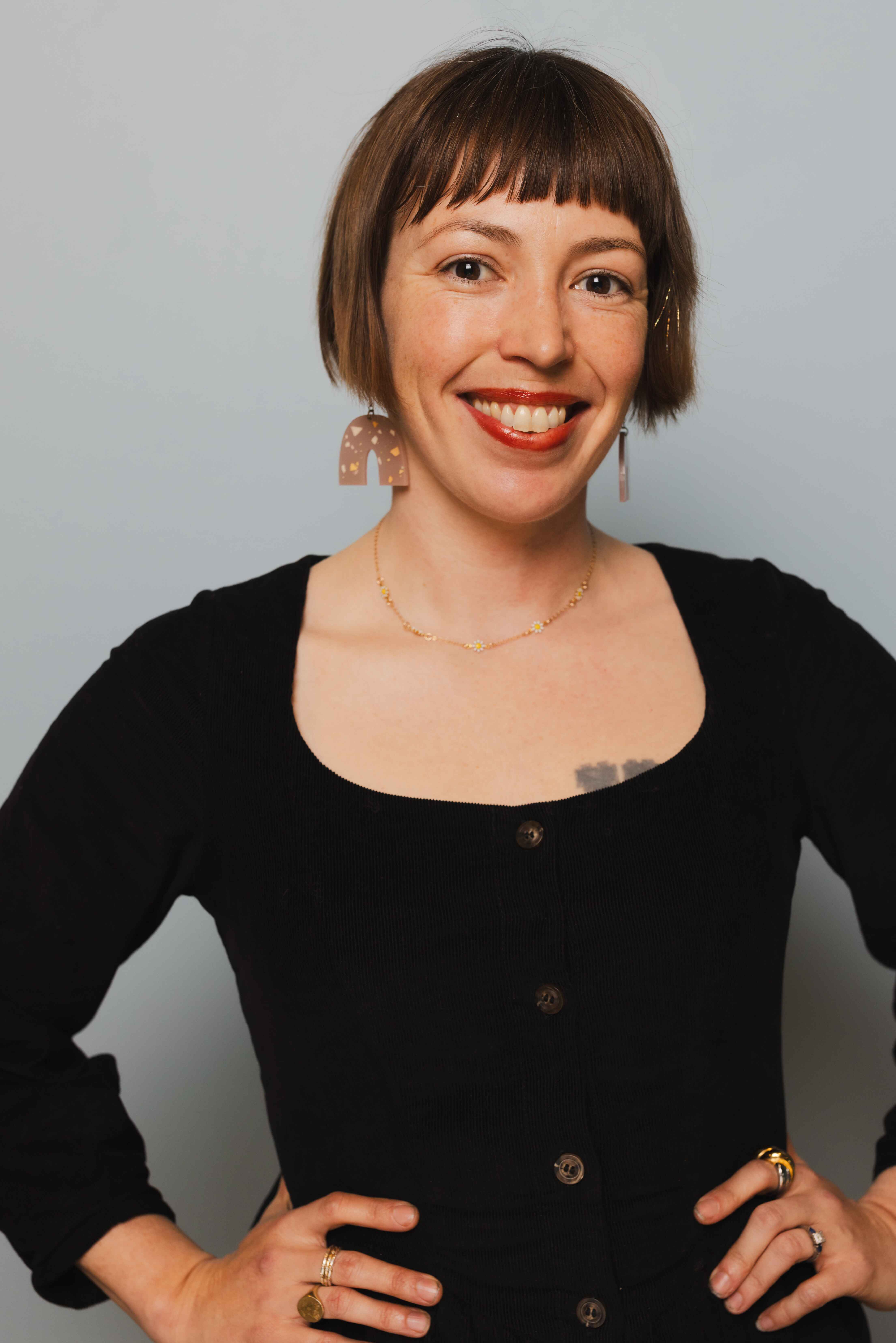 Headshot of Ashley Brewer, a white woman with short brown hair. She is wearing a black dress and smiling, with her hands on her hips.