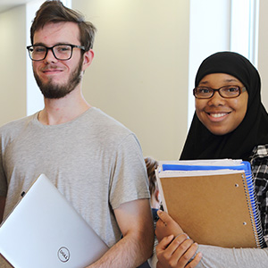 two students, one male one female smile while holding a computer and some notebooks