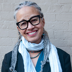 an image of a woman with braids wearing glasses with black frames and a blue scarf with a grey brick background