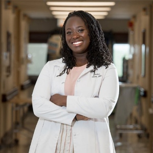 The image shows a black woman standing in a hospital hallway in her white coat smiling and looking at the camera with arms crossed in front of her