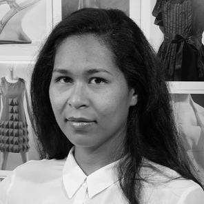 a woman looks at the camera with images of dresses behind her, her hair is dark and down and she is wearing a white shirt with a collar