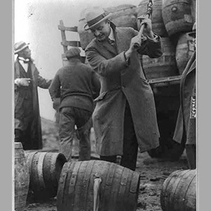 a black and white image of a man with an ax swung over his shoulder standing over a barrel with a hole in it and liquid pouring out.