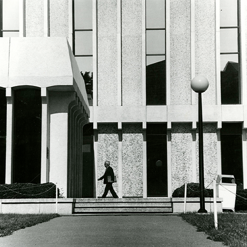 black and white image of the outisde of a concrete building with a man walking in front of it