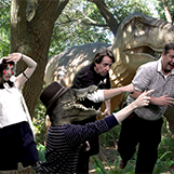 VCU Libraries employees pose in front of a green screen. The background show a T-rex. One employee is dressed as Princess Mononoke. Another is wearing a lizard mask and a fedora.