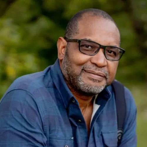 The image shows a black man with close shaven hair wearing black framed glasses and a blue button down shirt against a wooded background