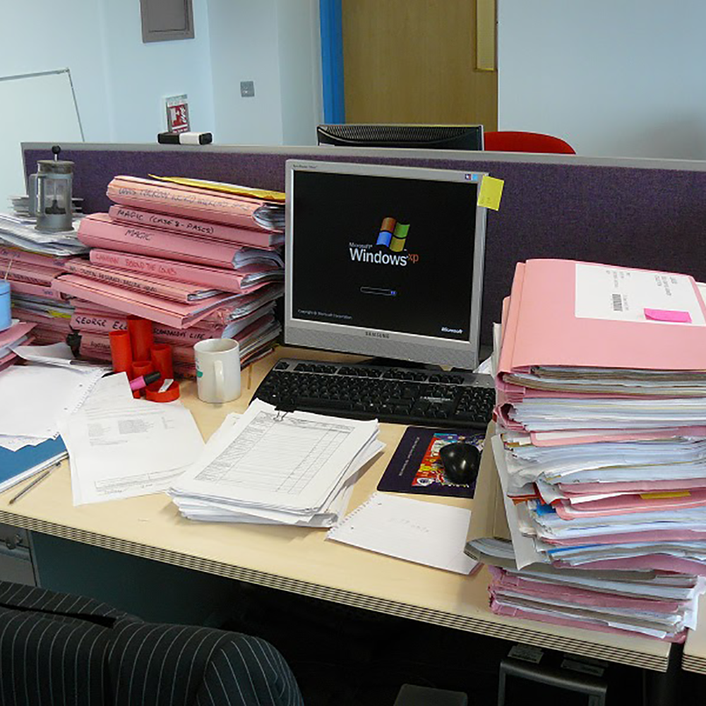 An office desk covered in pink file folders stuffed with papers. In the center of the desk is a computer monitor with a Windows XP startup screen.