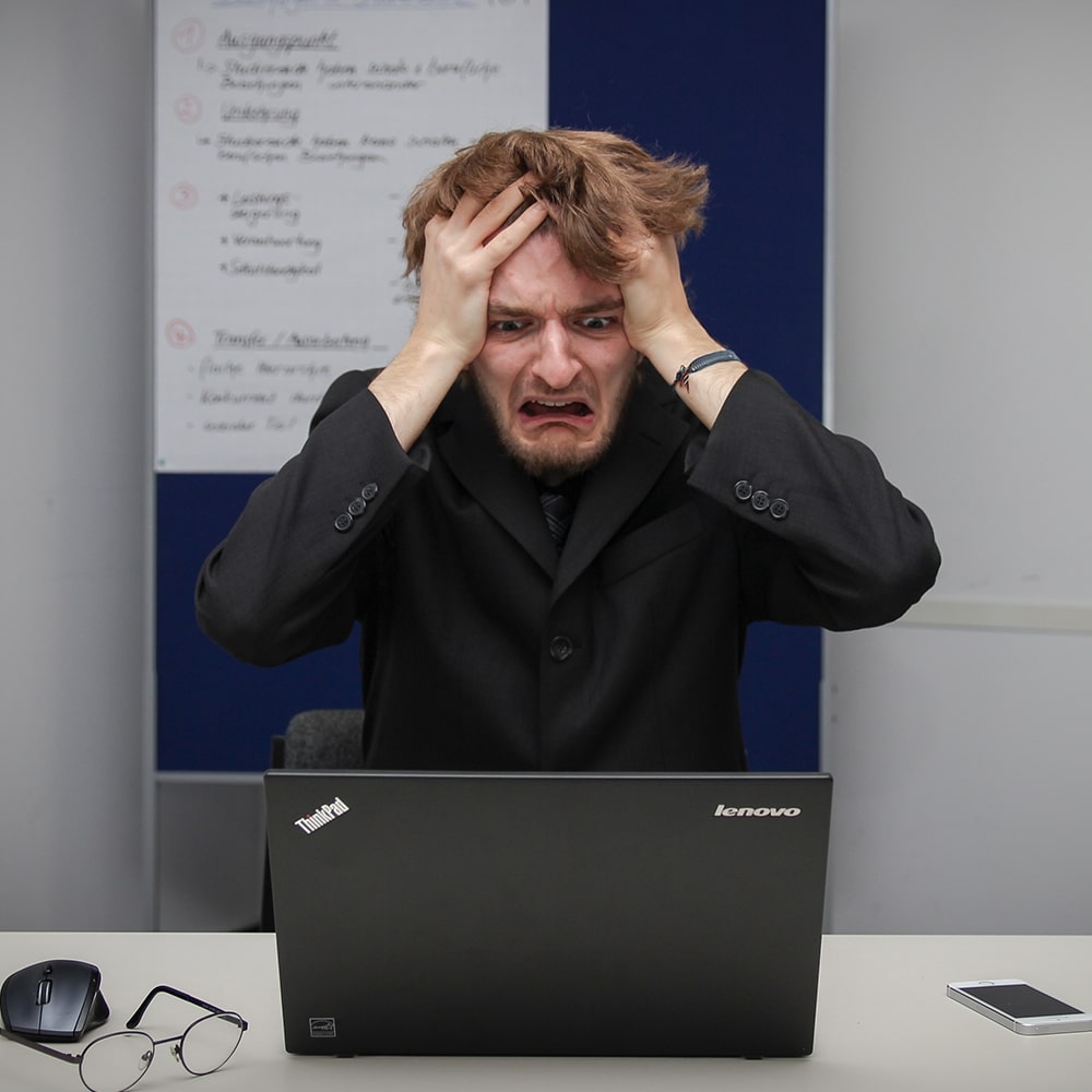 A man in a suit holding the sides of his head and looking in frustration/horror at a laptop screen