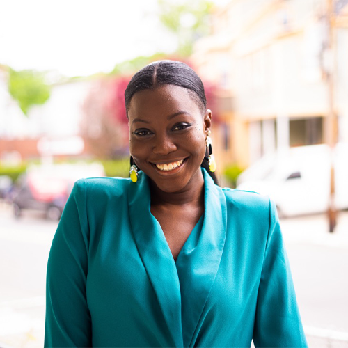 The image shows a black woman with hair slicked back wearing bright yellow earrings and a bright blue blazer smiling at the camera outdoors in front of a city street