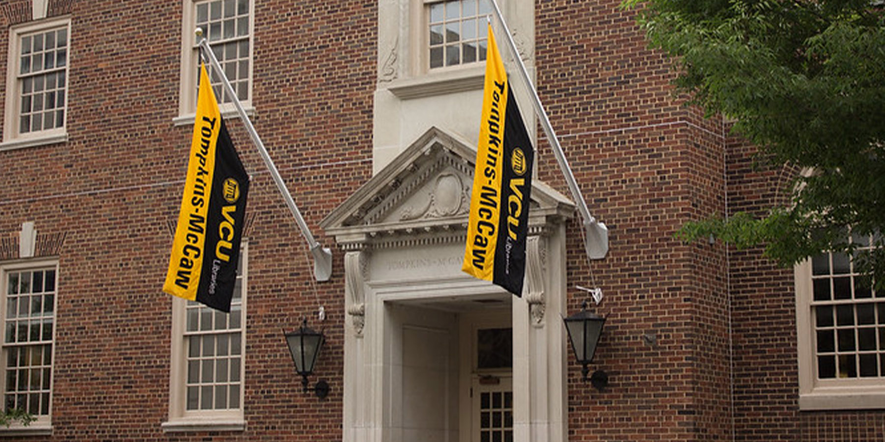 A photo of the front of the Medical College of Virginia campus library with the words Tompkins-McCaw Library engraved over the door with flags of black and gold with Tompkins-McCaw Library.