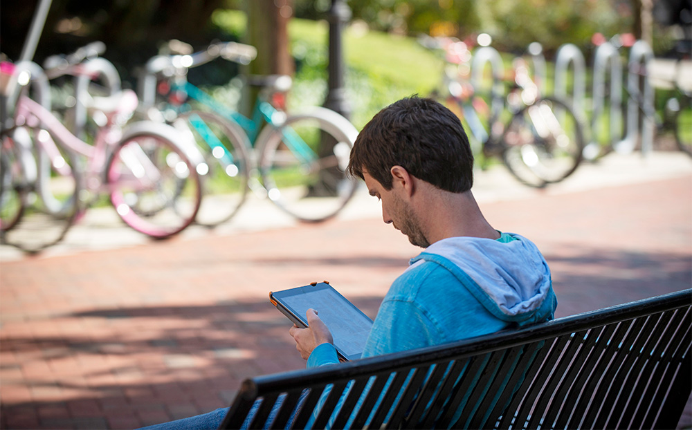 A young man sits on a bench reading from a tablet.