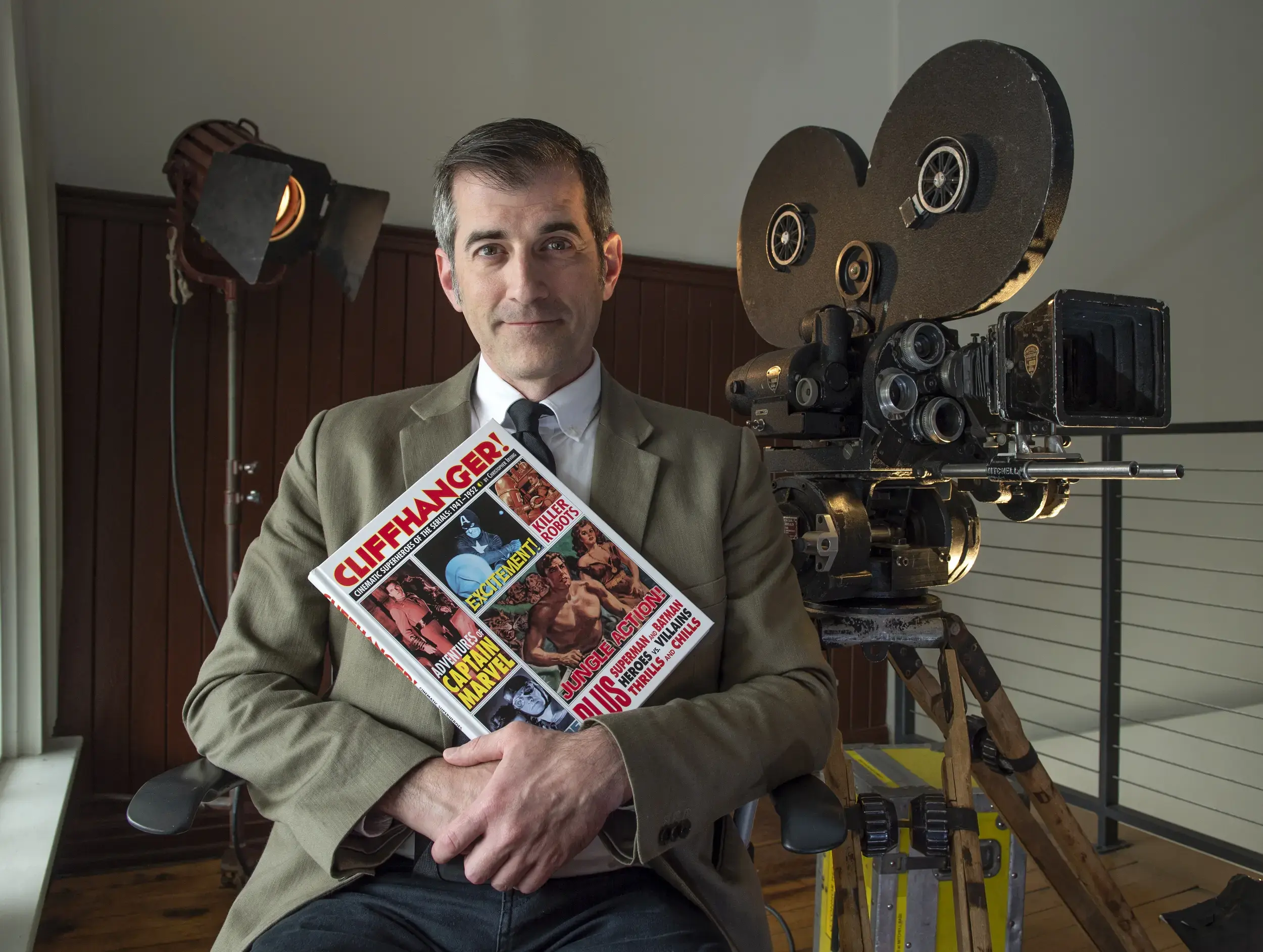 A man sitting in front of a film camera holding a book that says 