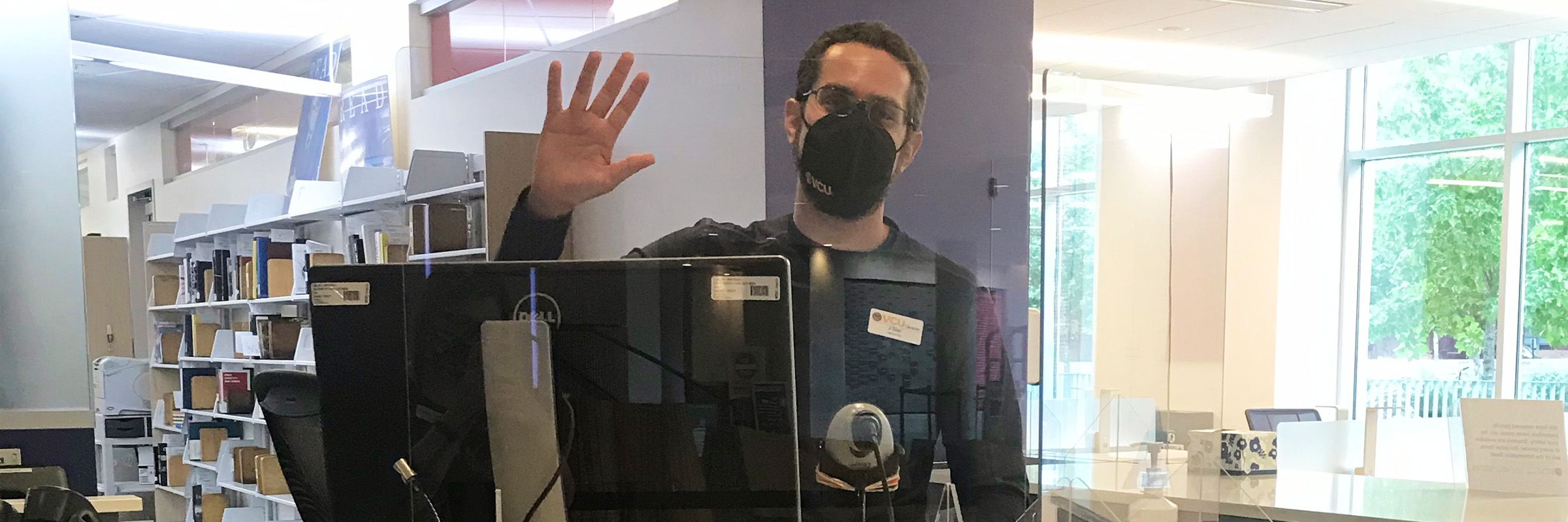 Library staff member, wearing a mask, waves behind a plexiglass shield at an information desk.