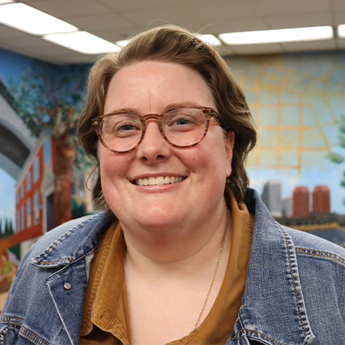Portrait of Kelsey Cheshire in front of a mural. Pronouns She/Her.