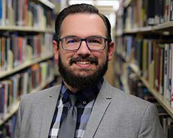 Portrait of Tim Siegel in front of book shelves. Pronouns He/His.